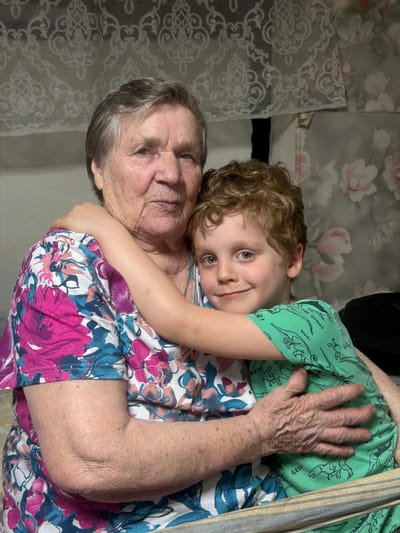 An elderly woman and a young child are closely embracing. The woman has gray hair and is wearing a floral-patterned dress in pink, blue, and white. The child, with light brown curly hair, is wearing a green shirt with dinosaur prints. The woman is holding the child close, with her arm and hand visible. The background includes floral wallpaper in shades of gray and pink, and patterned lace-like fabric. The image's orientation suggests it may have been rotated. The city of Dubrova, Russia is not identifiable from the image.
