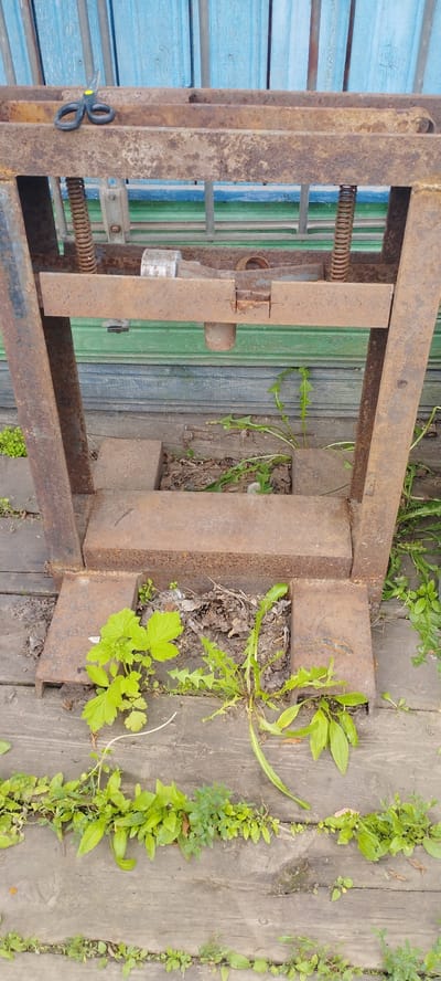 The image shows a rusty metal press machine outdoors, situated on a wooden platform. Green plants and weeds are growing around and through the base of the machine. In the background, there is a blue wooden fence. A pair of black scissors with yellow handles rests on the upper part of the machine. The environment suggests an outdoor, possibly rural or neglected setting. The location is specified as Pokosnoye, Russia.