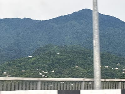 This is a wide-angle, horizontal, outdoor, daytime photograph taken from an elevated perspective, likely from a vehicle or a bridge, looking towards a lush, forested mountainside. A large, dark green mountain dominates the background, its slopes densely covered with trees, suggesting a temperate or subtropical climate. The sky is overcast and a pale grey. In the mid-ground, smaller hills are also covered in a similar dense forestation, with scattered white houses nestled among the trees. The houses vary in size and appear to be residential dwellings. In the foreground, a portion of a metal railing or barrier is visible, with vertical slats and horizontal capping. A tall, vertical metal pole, out of focus, bisects the mid-ground and background of the image on the right side. The overall impression is of a rural or semi-rural mountainous landscape near Chakvi, Georgia, characterized by abundant greenery and scattered human settlements.