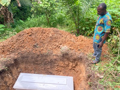 A Black man stands to the right of a freshly dug grave in a lush, green environment in Igbara, Nigeria. The grave is a deep rectangular pit in the earth, with a pile of reddish-brown soil to its left. A white, rectangular coffin is visible at the bottom of the grave, partially covered by plastic sheeting. The man, wearing a brightly patterned blue, yellow, and red shirt and grey pants, looks down into the grave. The surrounding area is dense with tropical vegetation, including palm trees and various leafy plants. The overall scene suggests a burial taking place.