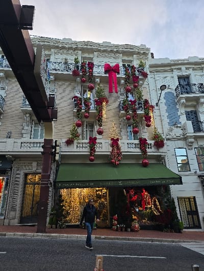 A decorated building facade is visible. Red ornaments, ribbons, and foliage adorn balconies and window ledges. A green awning covers part of the ground floor storefront. The storefront window is illuminated with golden lights and reflective surfaces. A man in a dark jacket is walking near the building. A street is to the side of the building.