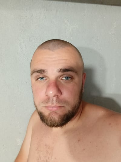 A bald man with blue eyes and a beard is photographed from the chest up, against a textured gray wall. His nose is red, and his skin appears tanned. He is shirtless, revealing some chest hair. The background is a plain, light gray wall with some subtle texture and slight discoloration. No other people or objects are visible in the image, and there is no indication of the location being Pishchane, Ukraine.