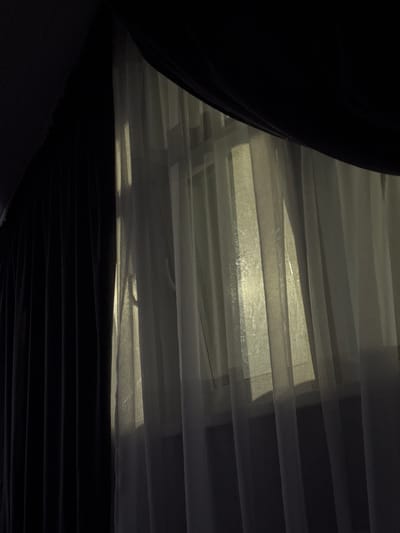 The image shows a close-up view of dark curtains on the left and sheer white curtains on the right, allowing light to filter through from behind. Vertical folds are visible in both sets of curtains. The lighting suggests it is either dawn or dusk, with soft, diffused sunlight casting subtle shadows through the sheer fabric. The environment appears to be indoors, possibly a residential setting, given the presence of curtains. There are no people or overt actions depicted in the scene. The location provided, "Doma 1096 Km, Russia," does not offer any visual cues within the image itself.
