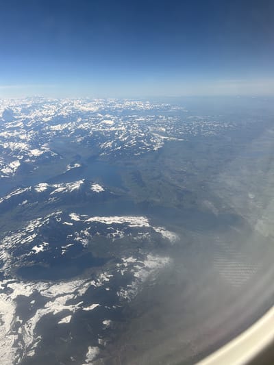 The image presents an aerial view captured from an aircraft window, looking down upon a vast, clear landscape. The upper half of the frame shows a wide expanse of bright blue sky. Below this, a sprawling mountainous terrain dominates the scene, characterized by numerous peaks and ridges with significant, patchy snow cover. Several large, irregularly shaped bodies of water, appearing as deep blue lakes, are nestled among the mountains and valleys. Extensive green and dark forested areas are visible between the snow-dusted elevations and surrounding the lakes, indicative of valleys and lower terrain. The lower right portion of the image reveals the curved white frame of an aircraft window, with atmospheric haze or condensation partially obscuring the view and displaying a faint ripple pattern on its surface. No individuals, specific man-made structures, or direct actions are discernible within the landscape. The scene depicts a high-altitude observation of a natural, rugged environment. The location is Peschiera Borromeo, Italy.