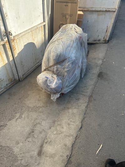 The image depicts an outdoor scene in the city of Kara-Suu, Kyrgyzstan. In the foreground, a large object wrapped in clear plastic sheeting is positioned on a concrete surface. The object appears to be a piece of equipment or furniture, possibly a lawnmower or a similar machine, with its form suggested by the bulky plastic wrap. Dark brown straps are visible, securing the plastic around the object. The concrete ground shows signs of wear and tear, with cracks and patches of discoloration.

To the left of the wrapped object is a large, weathered metal gate or door, also showing signs of rust and age. The gate is partially open, revealing a section of a white, textured surface behind it. Sunlight casts a shadow of an unseen object onto the gate.

In the background, to the right of the wrapped object, is a large, industrial-style metal bin, its surface weathered and stained. Part of a cardboard box, with some Russian text visible, is positioned on top of or next to the bin. The text on the box appears to be product information or branding.

The overall environment suggests an industrial or storage area, possibly a back alley or yard, characterized by worn surfaces, discarded materials, and utilitarian structures. There are no people directly visible in the image.