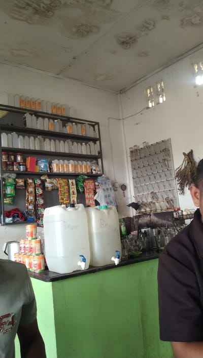 The image shows the interior of a small store or establishment in Meunasah Pupu, Indonesia. In the foreground, a person's shoulder and upper arm are visible on the right side of the frame. Two large white plastic water dispensers with blue spigots are placed on a counter in the center of the image; text that appears to read "Bitcoin" is written on each dispenser in black marker. To the left of the dispensers, a stack of canned goods is visible. The counter itself is a dark color, and it sits atop a bright green base.

Behind the counter, shelves are filled with various items. The top shelf holds rows of white and orange plastic bottles. The shelf below contains jars and boxes. The third shelf down is stocked with a variety of packaged goods, including what appear to be snacks and small sachets of products. A rack with numerous small compartments is mounted on the back wall, holding what seem to be glass bottles. To the right of this rack, a bunch of dried plants hangs. The ceiling has visible water stains. The lighting suggests it is daytime, with sunlight coming through a window on the right side of the room.