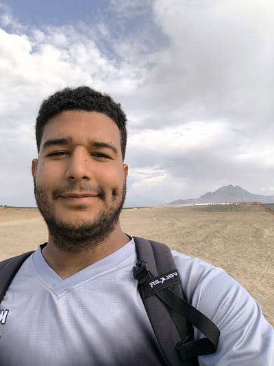 A medium shot captures a person with dark, curly hair, and a beard, smiling slightly with a visible dimple. The individual is wearing a light gray V-neck shirt and a black backpack with white lettering "REKJAV" visible on one of the straps. The setting appears to be a desert or beach environment, with a mountain visible in the distance. A low wooden fence runs along the flat expanse of sand, suggesting a boundary or pathway. The sky is overcast with a mix of white and gray clouds.
Based on the reference to Huanchaco, Peru, the location may be near or on the beaches of Huanchaco or potentially the surrounding desert areas near the coastline. The mountain in the background could be part of the local landscape.