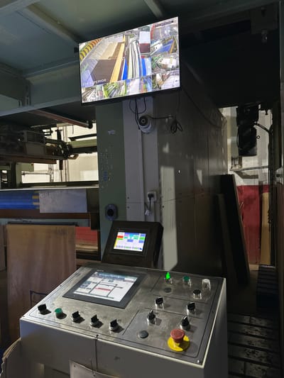 The image depicts an industrial setting, possibly a factory or manufacturing plant, location: Piñeyro, Argentina. The scene is dominated by heavy machinery, control panels with various buttons and displays, and a CCTV monitor showing multiple camera feeds of the production line. The lighting is dim, and the machinery appears aged and worn. There are no people visible in the immediate frame.