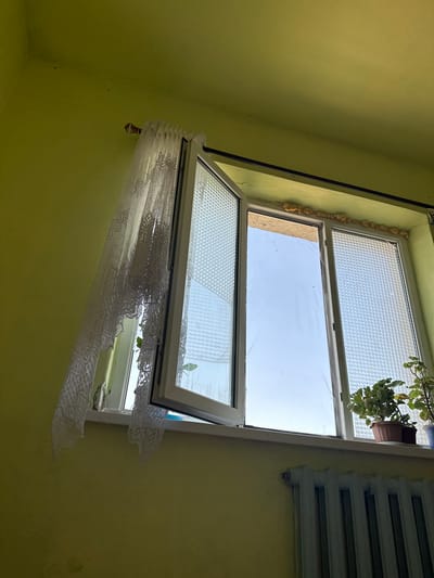 The image shows a close-up, low-angle view of an open window in a room with light green walls. A white lace curtain hangs on the left side of the window, partially obscuring the view outside and the textured glass of the window pane. The window itself appears to be a double-pane casement window, with one section swung open. The windowsill is white and holds two potted plants, one with green leaves in a brown pot and another partially visible plant in a blue pot. Below the windowsill, a section of a light gray radiator with multiple vertical columns is visible. The top edge of the window frame shows some yellow insulation foam. The sky visible through the open section of the window is a pale blue. The environment appears to be an indoor residential setting. The location within the city of Koçkor-Ata, Kyrgyzstan is not discernible from the image itself.