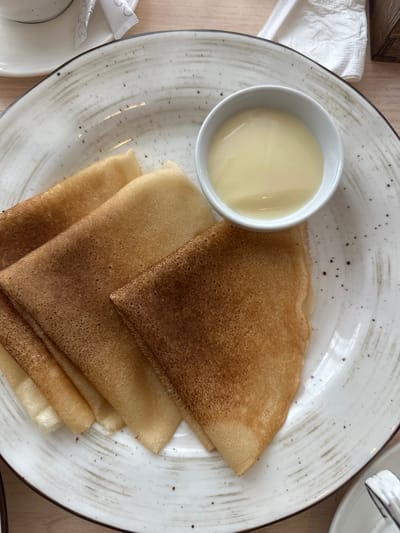 A close-up, overhead shot showcases a white ceramic plate with a textured, speckled design, holding several folded crepes and a small white dish of condensed milk. The crepes, varying in shades of golden brown and darker toasted edges, are neatly arranged in a fan shape. The condensed milk in the dish is a pale yellow, smooth, and creamy. The plate is positioned on a light-colored wooden surface, suggesting a table setting. The background is softly blurred, hinting at other elements of a meal or cafe environment, such as a white napkin and a partial view of a cup and saucer. The scene is lit from above, creating subtle highlights on the food and plate. The location is a cafe in Slyudyanka, Russia.