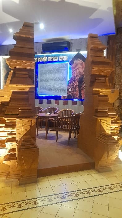 The image depicts an indoor scene within an establishment in Glantengan, Indonesia. In the foreground, a light-toned tiled floor with a dark, ornate border transitions to a raised, darker step. This step leads into an area framed by two large, light-brown, multi-tiered structures resembling traditional Indonesian architectural gate elements, positioned on the left and right. Centrally positioned beyond these structures, on a raised platform, is a circular wooden table with three matching wooden chairs arranged around it; an ashtray rests on the table. Behind the seating arrangement, a large, rectangular display panel is affixed to a wall. The panel features a textual description in Indonesian, including the phrase "BUDAYA MENARA KUDUS," and an image of a red-brick minaret, consistent with the Menara Kudus. Blue LED lighting borders the display panel. An air conditioning unit is mounted on the wall above the display. The ceiling, visible in the upper portion, contains recessed light fixtures. No individuals are present in the static scene.