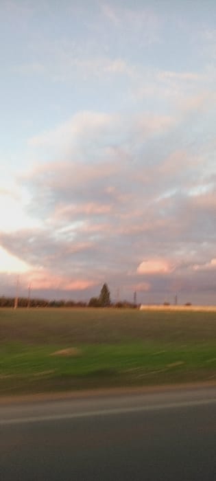 An orange-tinged sunset illuminates a sky filled with striated clouds. Below, a green field fades into indistinct foliage. A gray, possibly asphalt, surface occupies the extreme right. The image is blurry, particularly at the bottom. It appears to have been taken quickly, possibly from a moving vehicle.