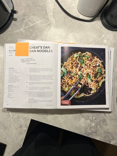 A cookbook is open to a recipe page for "Cheat's Dan Dan Noodles." The page includes an overhead photo of the finished dish: noodles, greens, and meat in a dark sauce, being stirred in a black pan with silver tongs. The recipe lists ingredients, including pork sausages, five-spice powder, pak choi, spring onions, dark soy sauce, chili oil, and straight-to-wok noodles. Instructions cover cooking the sausage meat, preparing the pak choi, making the Dan Dan sauce, and combining everything in the pan. A "Speed Hack" suggests using five-spice sausage meat to emulate the traditional Sichuan flavor.