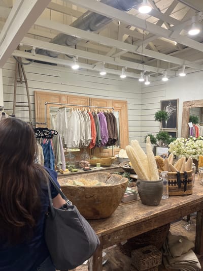 The image captures the interior of a retail store, likely a boutique, in Fredericksburg, Virginia, USA. Overhead, a white painted open ceiling structure with lights is visible. A rack of clothing hangs against the wall, displaying an assortment of garments in various colors and styles. A wooden frame surrounds the clothing rack. To the right, a wooden table displays an array of merchandise, including decorative items, brushes, and natural sponges. A person with long, brown hair, wearing a blue jacket and carrying a gray bag, stands in the foreground, appearing to be browsing or examining the items on display. The overall atmosphere suggests a curated, boutique shopping experience.