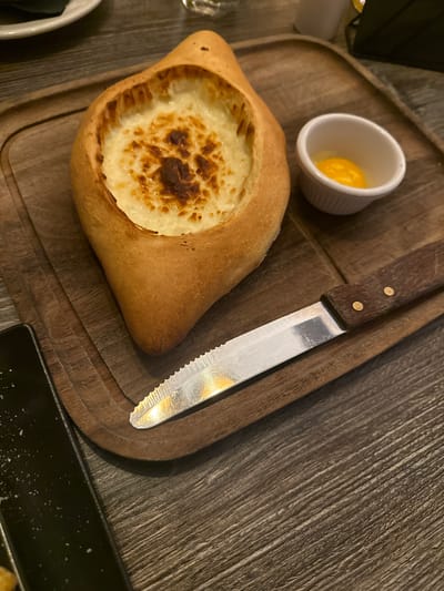 The image shows a Khachapuri, a traditional Georgian cheese-filled bread, served on a wooden board alongside a small white ramekin containing what appears to be an egg yolk and a knife. The Khachapuri is shaped like a boat with a filling of melted cheese, some parts of which are browned. The knife has a serrated blade and a wooden handle secured with rivets. The wooden board has rounded corners and edges. Part of a black plate is visible in the lower right corner. The table surface under the board has a textured, dark gray appearance, resembling wood grain. The location is implied to be Glenview, United States of America, based on instructions, but there are no specific visual cues to confirm this. There are no people visible in the image.