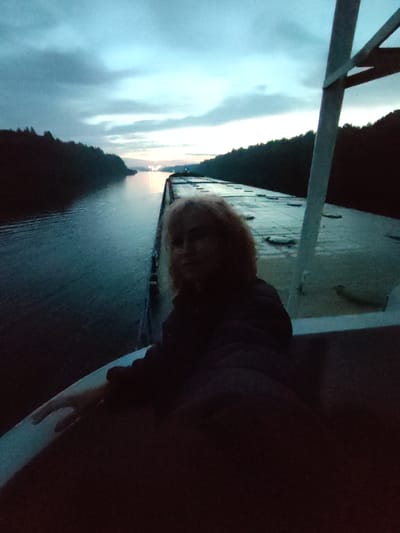 The image depicts a person with blonde hair looking at the camera from the deck of a vessel. The person is wearing dark clothing. The vessel appears to be in a body of water, possibly a river or canal, with tree-lined shores visible on both sides. The sky is overcast, with a hint of twilight or dawn. The scene is dark and somewhat blurry, suggesting it was taken in low light conditions. There is no specific indication of the location being within Annenskii Most, Russia. The image shows the stern of a vessel, with a railing and part of the deck visible. The water has ripples, indicating movement. The overall impression is of a serene, twilight setting on a waterway.
