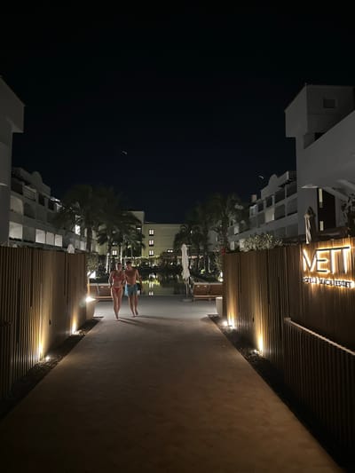 An exterior night shot captures a couple walking on a pathway towards a body of water. The pathway is lined with wooden slats and illuminated by warm lights. To the right, a sign reads "METT HOTEL & BEACH RESORT." Palm trees and white buildings with illuminated windows flank the scene, suggesting a resort or hotel environment in Playa Del Sol-villacana, Spain. The sky is dark and clear, with a faint streak of light visible in the upper portion of the image, possibly an airplane or satellite. The couple is walking side-by-side, appearing to be enjoying a relaxed evening stroll. The overall atmosphere is serene and inviting.