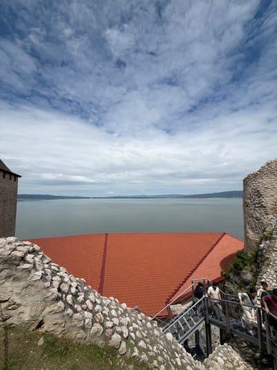 This is a high-angle, full-frame shot of the Golubac Fortress in Golubac Municipality, Serbia, on a cloudy day. The fortress is situated on a rocky outcrop overlooking a wide body of water, likely the Danube River. In the foreground, on the right, a section of a stone battlement is visible, with a modern metal staircase descending. Several people are on the stairs, walking up and down. A large section of a building with a red tile roof is prominent in the mid-ground. To the left, another stone battlement is visible, extending partially into the frame. The background features a vast expanse of water stretching to the horizon, with rolling hills and a hazy sky with scattered clouds. The overall scene depicts a historical landmark accessible to tourists.