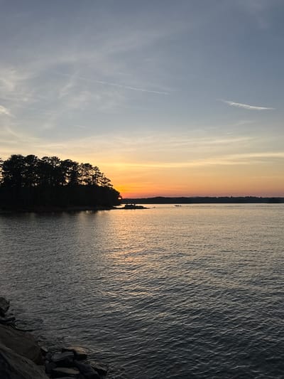 The image depicts a tranquil lake scene at sunset. A silhouetted treeline of coniferous trees forms a prominent feature on the left side of the frame, extending out into the water. The sky above displays a gradient of colors, transitioning from a soft blue at the top to a warm orange and yellow near the horizon, indicative of the setting sun. Wispy clouds are scattered across the sky, with a faint contrail visible in the upper portion.
The water in the foreground is calm, with gentle ripples reflecting the warm hues of the sunset. Rocks are visible along the left bank of the lake. In the distance, a treeline marks the far shore, and a small boat with two occupants can be seen on the water, creating a sense of scale and activity within the otherwise serene landscape.
This scene is consistent with locations around Lake Lanier, which is situated in the city of Sugar Hill, Georgia, United States. The presence of a large body of water, a silhouetted treeline, and the characteristic sunset colors are all common elements of this area.