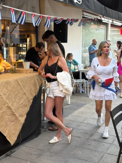The image depicts a scene in Cambrils, Spain. In the foreground, a table covered with burlap and a black cloth is partially visible. Two individuals are standing nearby, one wearing a black top and white shorts, the other in a black shirt. Cuban flags are hung on a storefront awning. In the background, a woman in a white dress with a red and blue sash is visible, along with other people sitting and standing in a street-side setting.