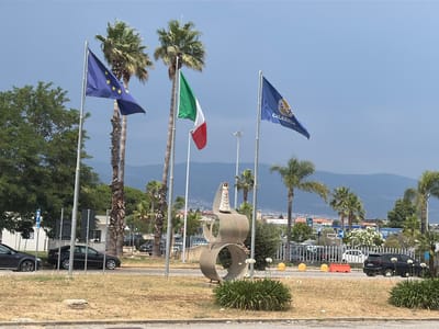 The image depicts a street scene in Lamezia Terme, Italy, featuring flags, a sculpture, and vehicles. In the foreground, a circular sculpture with a wave-like design stands on dry grass, adorned with white flowers. To the left, a blue European Union flag and a green, white, and red Italian flag fly on poles, with palm trees in the background. Further to the right, a blue flag displaying a crest and the word "CALABRIA" is visible. Several cars are parked along the roadside, including a dark sedan on the left and a black hatchback on the right. In the mid-ground, a white fence separates the road from other areas, with buildings and distant hills visible beyond. The sky is mostly clear with some clouds.