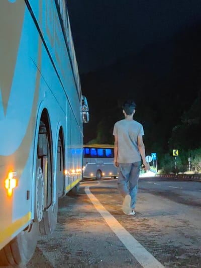 In the city of Sambal, India, at night, a person is seen walking away from the camera on the side of a road. The person is wearing a grey t-shirt and light-colored pants, with white shoes. They are holding a thin, metallic object in their right hand.
To the left of the person, a large, light-colored bus is parked, its side illuminated by amber lights. The bus has blue-tinted windows and large wheels. Further down the road, another bus is partially visible, also with illuminated blue windows.
The road has white markings and is dark, with trees and foliage visible in the background. Traffic signs, including a blue sign with a U-turn symbol and a yellow sign with a curved arrow, are present on the right side of the road. The overall scene suggests a roadside location at night, possibly near a bus stop or rest area.