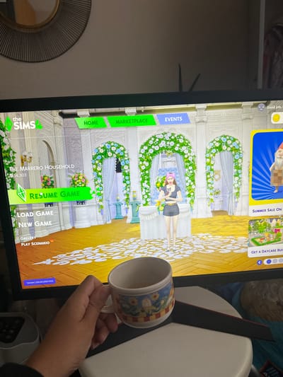 The image shows a person's hand holding a mug, in front of a computer screen displaying the game "The Sims 4". The game's main menu is visible, with options like "Resume Game", "Load Game", and "New Game". The background of the game appears to be a wedding or event setup with floral decorations. A character from the game, a woman dressed in a black outfit, is also visible.
The screen also displays other game-related information such as "The Marrero Household" with a sum of "$14,303", and various tabs like "Home", "Marketplace", and "Events". In the upper corner of the screen, there are notifications or promotions related to a "Summer Sale" and "Get a Daycare Build". The game version "1.124.63.1020 DX11" is also displayed.
The mug held by the person has a patterned design with flowers and what appears to be a gnome or a similar character. The overall scene suggests a casual setting, possibly a living room or office, where someone is taking a break from playing a video game. The lighting in the image is not strong, suggesting it could be daytime or the room lighting is subdued. There are no obvious weather cues. The location context of Macclesfield, United Kingdom, does not provide any specific visual cues within the image itself. The overall mood seems relaxed, as indicated by the person's posture and the activity of playing a game.