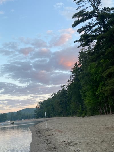 The image depicts a serene beach scene at Parc Plage Pop, Duhamel, Canada, during either sunrise or sunset. The sky is a pale blue with scattered clouds tinged with pink and orange hues. To the right, a dense line of evergreen trees forms a backdrop, with some branches extending towards the sky. The beach itself is composed of light-colored sand, with some scattered debris and footprints. Gentle waves lap at the sandy shore, and the water appears calm. In the distance, across the water, there are faint outlines of hills and more trees. There are a few people visible on the beach in the background, along with some boats and structures near the water's edge, suggesting a recreational area. The overall atmosphere is peaceful and natural.