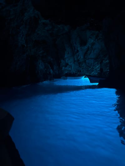 In the city of Komiža, Croatia, within the Blue Cave on the island of Biševo, a boat navigates the illuminated, crystal-clear water. The cave's interior is a spectacle of light and shadow, with the water glowing an ethereal blue due to sunlight filtering through an underwater opening. The rough, dark cave walls rise up on either side, contrasting with the vivid blue of the sea. A white tourist boat, with some passengers visible, moves through the water, its hull reflecting the brilliant blue light. Another similar boat is docked further in the cave. The scene is serene and otherworldly, capturing the unique natural phenomenon of the Blue Cave.