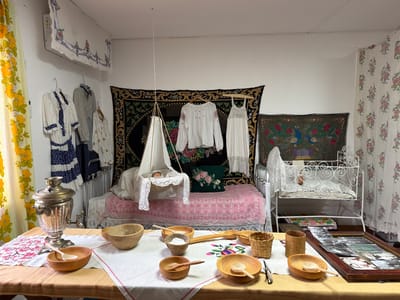 This image depicts an indoor display, likely in a museum or cultural center, showcasing elements of traditional life. The scene is set in a room with white walls and is filled with various objects. In the foreground, a table is covered with a light-colored tablecloth, adorned with embroidered patterns. On this table, there is a large, ornate samovar, several wooden bowls of varying sizes, and wooden spoons. To the right, a framed collection of old photographs is laid out.
The background features a bed with a pink and white patterned bedspread. Suspended above the bed is a white bassinet with a doll or baby figure inside. Clothing items, including what appear to be traditional embroidered blouses and dresses, are hung on a rack and on hangers. Decorative tapestries with intricate designs, including one with peacocks, hang on the walls. A white metal crib with ornate detailing is positioned to the right of the bed. Floral patterned curtains are visible on the left and right sides of the room. The overall impression is that of a staged exhibit, possibly recreating a historical domestic setting. The image is not specific to Ust'-Pyzas, Russia, as no identifying markers are present within the frame to confirm its location.