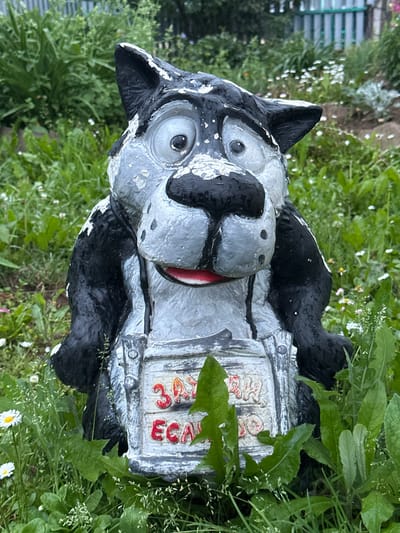 This is a close-up, eye-level, outdoor photograph of a weathered, cartoonish statue of a wolf in a garden setting. The statue is predominantly black and gray with peeling paint, and it is holding a sign with red Cyrillic lettering that reads "ЗАЯЦ И ЕСАУЛ" (Hare and Captain). The wolf statue has large, round eyes, a black nose with visible nostrils, and a red mouth. It appears to be wearing a dark vest or coat. The statue is positioned in the center of the frame, surrounded by lush green foliage and small white wildflowers. In the background, a blurred green fence and more dense greenery are visible. The lighting suggests it is daytime. The context of Mel'nichnaya, Russia, is not visually discernible from the image itself.