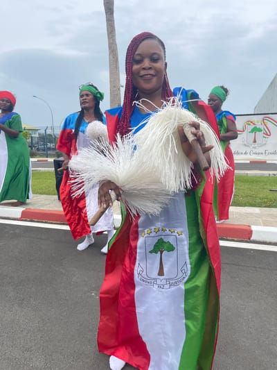 This image depicts an outdoor scene featuring several women participating in what appears to be a national celebration or parade in Malabo, Equatorial Guinea. The foreground shows a smiling woman with braided reddish-brown hair, looking directly at the camera. She is dressed in a blue top with vibrant red and green fabric draped around her, embodying the colors of the Equatorial Guinean flag. In her hands, she holds a large, white, fluffy ceremonial object, possibly a traditional whisk or rattle.

Behind her, other women are partially visible, similarly adorned with garments reflecting the national flag's colors and also holding white fluffy items. The national flag of Equatorial Guinea is prominent; its design includes horizontal green, white, and red stripes with a blue triangle on the hoist side, and the national coat of arms in the center. The coat of arms features a silk-cotton tree (ceiba), six yellow stars above it, and the motto "UNIDAD PAZ JUSTICIA" (Unity, Peace, Justice). This motto is clearly visible on the flags and a large emblem on a modern building in the background. The building's emblem also bears the full text "REPÚBLICA DE GUINEA ECUATORIAL."

The setting includes a paved road or public square, with green grass and palm trees on the left. The sky is overcast, indicating daytime. The joyful expressions of the participants, particularly the smiling woman in the foreground, convey a sense of national pride and festive spirit.