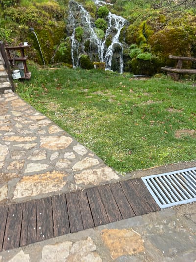 The scene depicts an outdoor area in Despotovac Municipality, Serbia, featuring a developed natural water source. In the background, a waterfall cascades down a steep, moss-covered rock face abundant with green vegetation. To the left, a dark spigot integrated into the rock emits a continuous stream of water. A white ceramic mug rests on the top tier of a dark, multi-tiered wooden stand positioned next to the spigot; a white bucket is visible on a lower shelf of the stand. The midground features a green lawn uniformly speckled with numerous small white particles, consistent with hail or fallen blossoms. To the right, a rustic wooden bench constructed from logs is situated near the base of the waterfall. The foreground consists of a path paved with irregular light brown and beige stones set in grey mortar. This stone path transitions to a section of dark wooden planks, adjacent to a rectangular metal grate embedded as a drain cover. The ground and surfaces appear damp, suggesting recent precipitation. No individuals are present in the image.