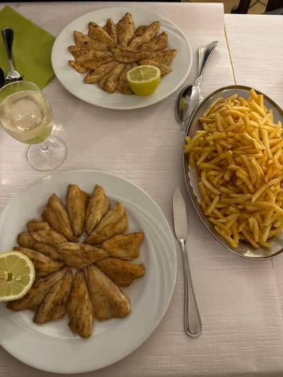 A table is set with two plates of what appears to be fried fish fillets arranged in a circular pattern, each garnished with a lemon wedge. A large silver platter filled with french fries sits on the table, alongside a butter knife and two spoons. One plate of fish sits next to a green napkin and a wine glass filled with a clear liquid. The table is covered with a white tablecloth. It is plausible this is a restaurant in Chêne-Bourg, Switzerland. No people are visible in the image.