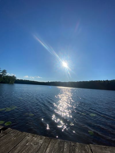 A bright sun shines in a clear blue sky over a calm lake, casting shimmering reflections on the water's surface. The sun's rays create a starburst effect with a rainbow halo. A dense line of dark green trees forms the horizon along the far side of the lake, with a few scattered white clouds in the sky. In the foreground, a wooden dock or pier extends into the water, showing weathered planks with visible grain and nail holes. Lily pads float on the dark blue water near the dock. The overall scene depicts a serene natural environment. The location is described as the Азёрскі сельскі Савет in Belarus. There are no people or animals visible in the image.