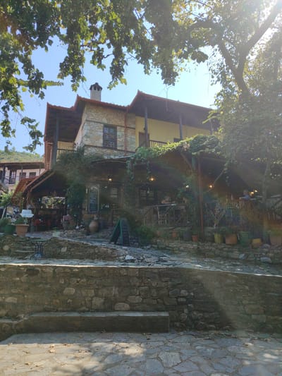 The image depicts a charming outdoor dining area in Palaios Panteleimonas, Greece. The scene is dominated by a traditional stone building with a wooden-tiled roof, featuring a second-story balcony and a ground-level veranda adorned with lush greenery. Several people are visible seated at tables, enjoying meals and drinks in the relaxed atmosphere.
The time of day appears to be late afternoon, suggested by the warm sunlight casting shadows and creating sunbeams filtering through the trees. The weather seems pleasant and clear, with a bright blue sky visible above the building and foliage. The setting is rustic and inviting, with stone walls, cobblestone pathways, and numerous potted plants adding to the ambiance. Hanging lamps on the veranda provide a warm glow, hinting at the possibility of evening dining.
Notable details include a stone chimney on the building's roof, several chalkboards displaying menus or information, and large terracotta pots scattered around the area. The architectural style is characteristic of traditional Greek villages. The overall mood is one of tranquility and leisurely enjoyment.
There is visible text on the chalkboards in front of the building.