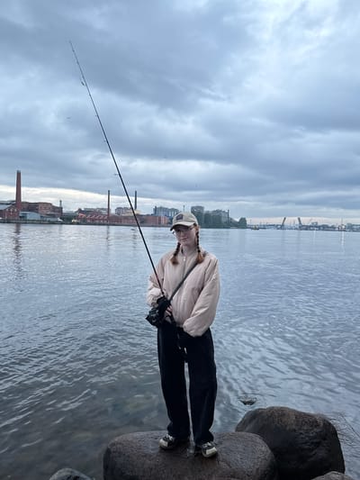 A young person with red braided hair, wearing glasses, a tan jacket, black pants, and a tan baseball cap, stands on large rocks by a body of water, holding a fishing rod. In the background, across the water, is the cityscape of Ladozhskii Trudposelok, Russia, featuring industrial buildings with red brick chimneys and modern structures. The sky is overcast with heavy clouds.