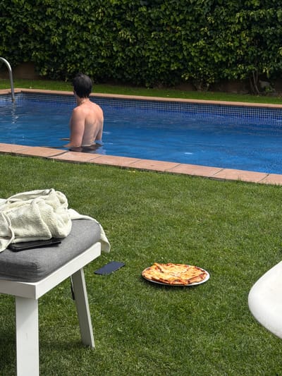A man is entering a swimming pool, which is bordered by a hedge on one side and a grassy area on the other. A pizza on a white plate sits on the grass near a white chair, which has a gray cushion and a white towel. A dark object, possibly a tablet or phone, is also on the grass near the chair. The scene appears to be in Sant Andreu de Llavaneres, Spain.