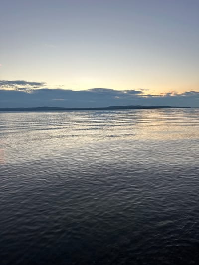 The image shows a vast body of water with gentle ripples reflecting the soft light of a twilight sky. In the distance, a low-lying landmass or island with a dark silhouette is visible on the horizon. Above the water, the sky transitions from a pale blue at the top to a warm gradient of yellow and orange near the horizon, where scattered clouds are present. The overall impression is one of tranquility and a serene natural landscape. The location is described as Горы, Russia.