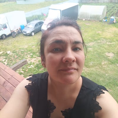This media file is a selfie taken outdoors in Podlubovo, Russia. The subject is a woman with dark hair and brown eyes, wearing a black top with lace detailing on the shoulders. She is looking directly at the camera with a neutral expression.
The background shows a grassy yard with several objects. To the left, there is a white truck and a motorcycle. A gray car is parked nearby. In the center of the background, there is a greenhouse with a transparent cover. Further in the background, a wooden fence runs along the edge of the property. The scene appears to be set during the daytime, with overcast weather suggested by the soft lighting. The overall setting suggests a rural or suburban environment. There is no visible text in the image.