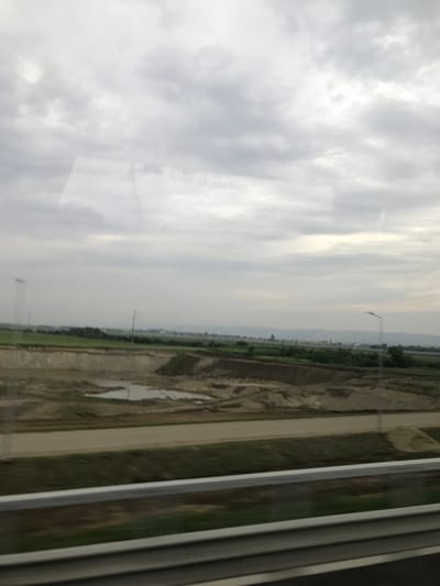 This forensic image captures an exterior view from the window of a moving vehicle, likely a train or car, presenting a rural or semi-rural landscape under a heavily overcast sky. The scene is located in the vicinity of Adjud, Romania.
In the immediate foreground, a multi-tiered metal guardrail, featuring a prominent light-colored top rail and two darker grey lower rails, horizontally spans the lower portion of the frame. Below the guardrail, a narrow, blurred strip of green vegetation is visible, consistent with a roadside or railway verge.
The midground is dominated by an extensive area of disturbed, excavated earth or sand, appearing in various shades of brown and tan. Within this area, several irregularly shaped puddles of standing water reflect the grey sky. A flattened, unpaved track or road of lighter brown color runs horizontally through the earthworks, separating them from cultivated or wild green fields beyond. Scattered utility poles with visible lines are interspersed throughout the midground and background.
In the background, a expanse of green fields or low vegetation stretches towards a distant, hazy horizon line, where a faint silhouette of hills or mountains is discernible. Indistinct white structures, possibly buildings or industrial elements, are barely visible against the distant landscape. On the far right, a street lamp with a diagonally extending arm is present.
The sky is entirely covered with thick, grey clouds, indicating an overcast day without direct sunlight. No people are visible in the scene. Faint, reversed textual reflections, including fragments like "JEU DE RECLAM" and numbers, are visible on the vehicle's window in the upper left and central portions of the image, obscuring parts of the sky. The overall impression is one of a vast, open landscape with signs of ongoing land development or agriculture, viewed from a moving transport route.