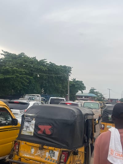 The image depicts a street in Alasia, Nigeria, congested with vehicular traffic. Predominantly yellow 'keke' (auto-rickshaws) are interspersed among other cars, including white sedans and minivans. The vehicles are in a standstill, suggesting a traffic jam. Lush green trees flank the road, partially obscuring buildings in the background. Utility poles with wires are visible. The sky is overcast. A person is partially visible in the foreground with a branded towel over the shoulder. Advertisements are affixed to the rear of one of the 'keke'.