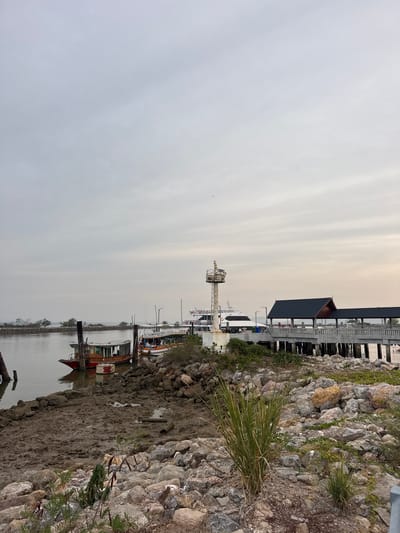The image shows a marina scene in Pak Nam Chumphon, Thailand on an overcast day. The foreground contains a rocky shoreline and muddy bank. In the middle ground, there are several boats moored in the water. One boat has a red hull. A lighthouse is visible in the distance, and behind it, a building with a dark roof and light-colored supports is present. The sky is a uniform grey with subtle striations.