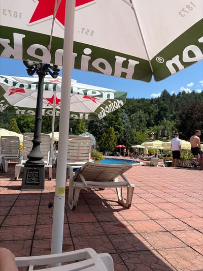 The image depicts an outdoor swimming pool area on a sunny day, with a focus on a large umbrella advertising "Heineken". The umbrella is white with a green band and red stars, the branding partially visible. In the foreground, there are red tiled paving stones and several white plastic lounge chairs, some casting shadows. A black decorative lamp post stands near the center. In the background, beyond the pool, there are more lounge chairs, yellow umbrellas, and a dense forest of green trees under a clear blue sky. Several people are visible near the pool: some are walking, others are standing by the edge, and two shirtless men are standing on a raised tiled area to the right, one leaning against a decorative fence. The location is identified as Zverino, Bulgaria.