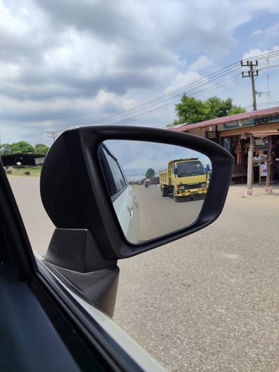 In Meunasahmee, Indonesia, a side-view mirror of a white car reflects a yellow dump truck driving on a paved road under a cloudy sky. The truck has the name "MITSUBISHI" on its front. In the reflection, other vehicles and a few pedestrians are visible in the distance. To the right of the car, buildings with red roofs line the road, and power lines are strung across the sky.