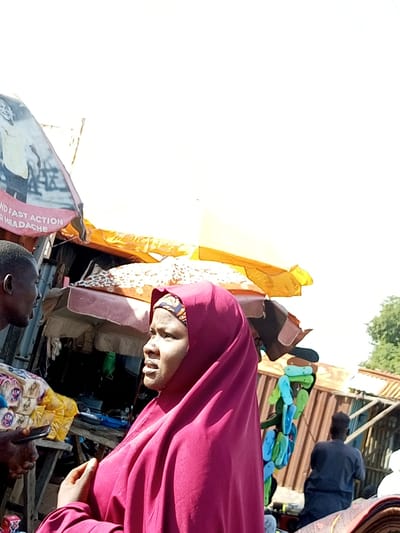 The image captures an outdoor marketplace scene in Gadao, Nigeria. In the foreground, a woman wearing a pink hijab is positioned in profile, facing right. To her left, a man is also shown in profile, looking towards her. Between them, there are several market stalls with various goods, including packaged items and what appear to be fabrics. Overhead, colorful awnings, one yellow and one orange, are visible. In the background, a man wearing a dark blue outfit is seated, facing away from the camera. Further in the background, more market structures and greenery are apparent. The lighting suggests it is daytime.