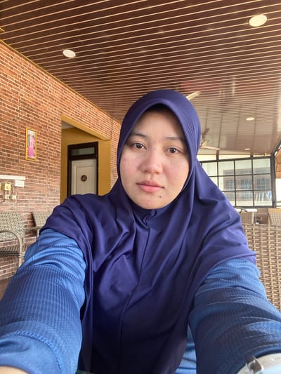 A selfie captures a young woman wearing a navy blue hijab and a textured blue long-sleeved shirt. She is seated outdoors, with a brick wall and a door visible behind her on the left. The ceiling above is made of wooden panels with visible light fixtures and a fan. Wicker chairs and tables are present in the background, suggesting an outdoor seating area, possibly a cafe or restaurant. The overall lighting indicates it is daytime. The location is identified as Keraton, Indonesia.