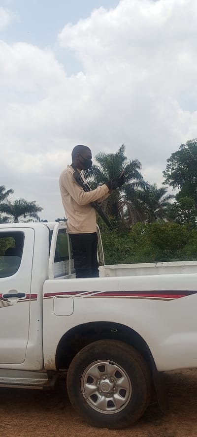 The image captures a daytime scene in what appears to be a rural or semi-rural area of Otunade, Nigeria. The central subject is a man standing in the bed of a white pickup truck, identifiable as a Toyota Hilux by the emblem on the wheel hubcap. He is wearing a light-colored long-sleeved shirt, dark trousers, and a black face mask covering his mouth and nose. The man is holding a dark long gun, resembling a rifle or shotgun, which is slung across his body. His gaze is directed downwards, suggesting he is looking at something held in his hands, possibly a mobile phone or another device.

The pickup truck is white with distinctive red and gold/brown striped decals running along its side. The ground beneath the vehicle appears unpaved, consistent with a dirt road or track. The background is dominated by lush green vegetation, including tall palm trees and dense foliage, indicating a tropical or subtropical environment. The sky above is bright and partly cloudy with large white cumulus clouds.

The activity depicted suggests a security presence, a patrol, or an escort, given the armed individual positioned in the back of the vehicle in an outdoor setting. The time of day is clearly daylight, likely mid-morning or early afternoon, with ample natural light and no harsh shadows. The man's expression is not visible due to the mask, but his posture appears calm and focused. No other individuals or specific events are clearly discernible in the frame.
