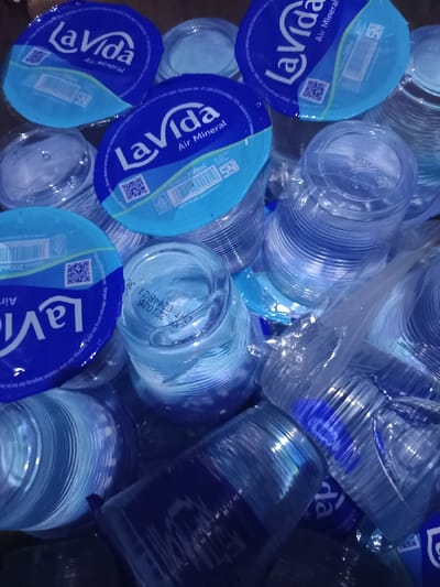 The image displays a cluttered pile of numerous individual servings of LaVida Air Mineral bottled water in clear plastic cups, situated in Limbangan Sari, Indonesia. Each cup, containing 200ml of water, is sealed with a circular foil lid that is predominantly blue with a light blue wavy design. The lids clearly feature the "LaVida Air Mineral" brand name, the volume "200ml," "SNI" certification, and QR codes. The cups are stacked and scattered haphazardly, some upright, others inverted or lying on their sides. One visible cup displays an expiry date of "EXP 221025 DNT 0244821 3" molded into its plastic rim. No individuals, actions, or interactions are discernible within the frame. The environment appears to be an indoor setting with a dark, indistinct background.