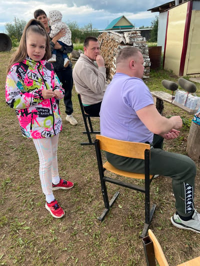 The image shows a group of people in what appears to be a rural or outdoor setting. A young girl is in the foreground, wearing a brightly colored jacket and red shoes. Behind her, several adults are seated, possibly on chairs or benches. One man is wearing a purple shirt and green pants with a distinctive snake-like pattern on the side. Wood and buildings are in the background, suggesting a countryside or village environment. Based on the location, this scene could be taking place in Большие Кусты, Russia.